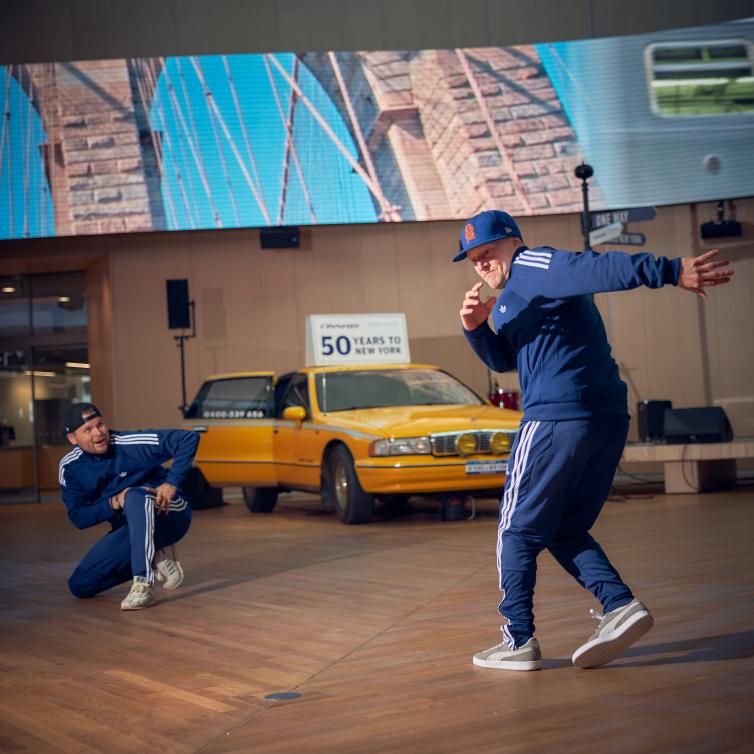 Kaksi veryttelyasuun pukeutunutta katutanssijaa tanssimassa taksin edessä.