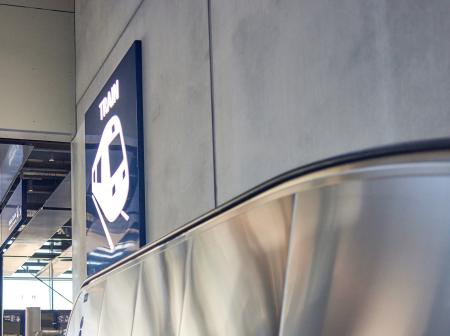 Juna-aseman kyltti harmaalla seinällä, liukuportaiden yläpuolella.