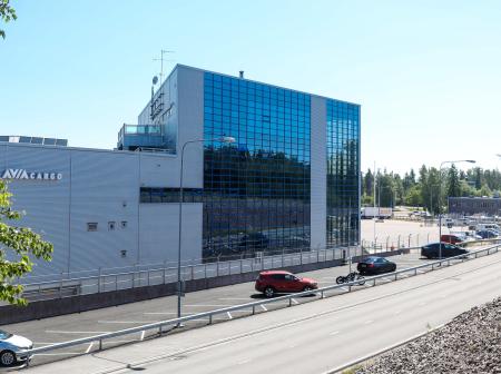 Kuvassa iso vaalea rakennus, jossa on tummaa lasiseinää ja parkkipaikka rakennuksen edustalla.
