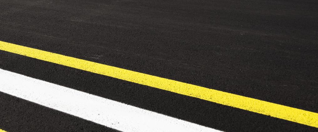 Lähikuva mustasta kiitotieasfaltista, jossa on keltainen ja valkoinen viiva.
