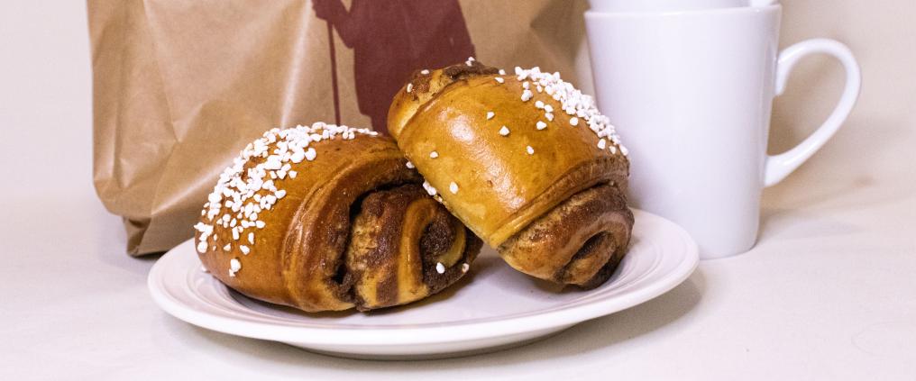 Kuvassa kahvimukeja, pullaa ja paperipussi.