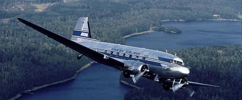 Vanha lentokone lentää metsän ja järven ylitse.