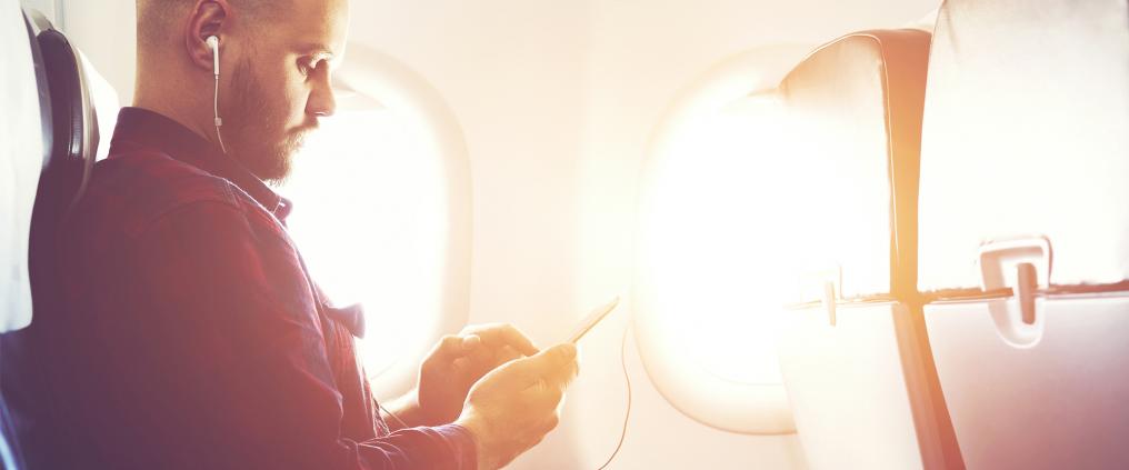Sun shines inside airplane cabin as passenger sits with earbuds on checking his phone.