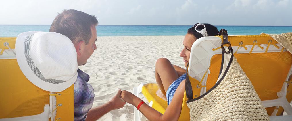 Pariskunta istuu aurinkotuoleilla rannalla ja pitävät toistensa kädestä kiinni.
