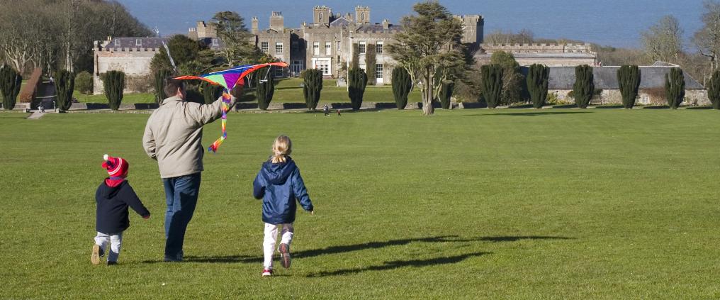 Father and two children are about to fly a kite in grasslands with a castle in the background.