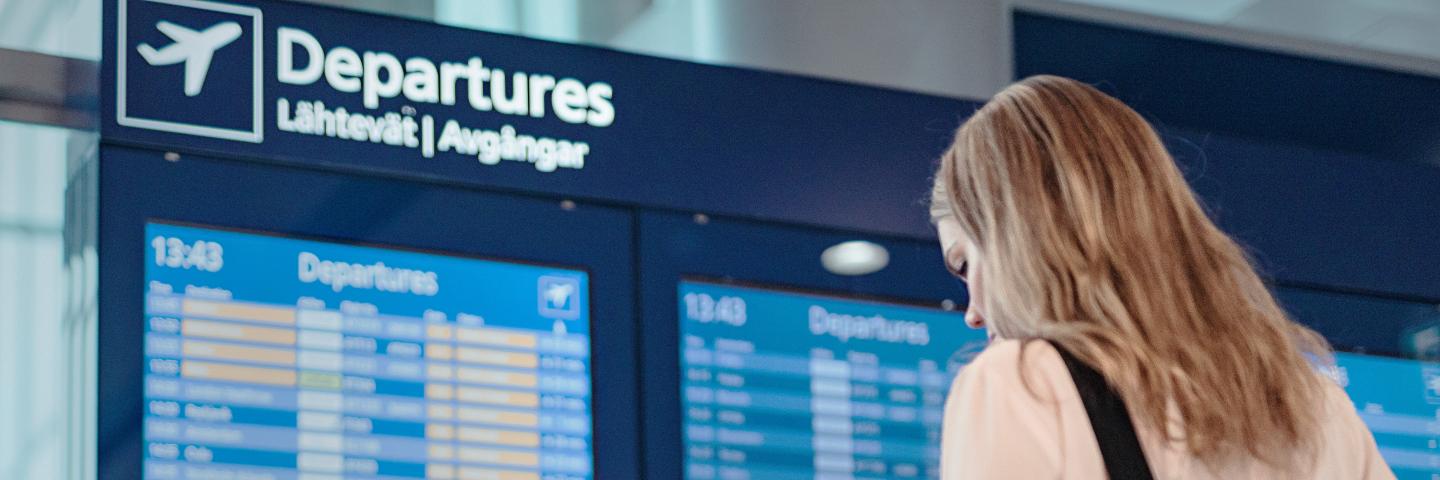 Matkustaja katsoo puhelinta lentotietotaulun edessä.