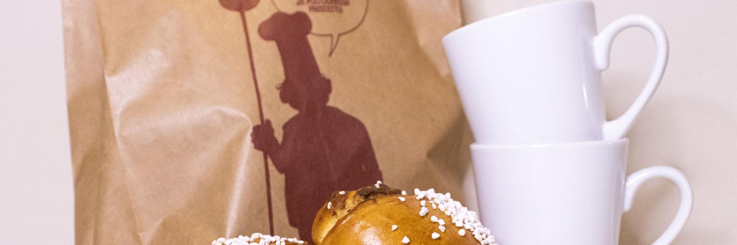 Kuvassa kahvimukeja, pullaa ja paperipussi.